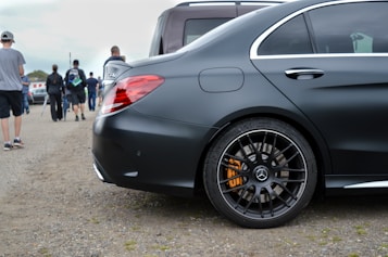 A sleek black car with tinted windows and a luxury brand emblem is parked on a gravel surface. People are walking in the background, some carrying bags, suggesting a public event or gathering.