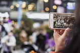 A person holding a smartphone taking a video of a group of people dancing in an outdoor, urban setting. The background appears blurred with bokeh lights, indicating a lively environment.