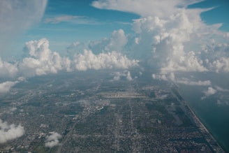 An aerial view of an urban area extending towards the horizon with numerous buildings, roads, and patches of greenery. Large, fluffy white clouds dominate the sky, creating a contrast with the blue sky visible above. The right side of the image features a coastline with water meeting the land.