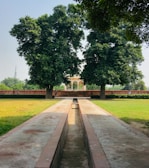 A beautifully landscaped garden with a long, narrow water channel leading towards a heritage structure framed by two large trees. The setting is serene, surrounded by grass and a few people walking along a brick pathway next to the water channel.