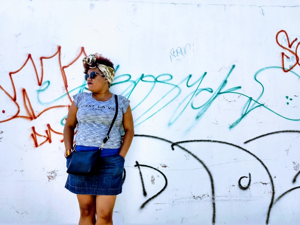 グレーのクルーネックのキャップスリーブシャツとブルーデニムのミニスカートを着た女性が、昼間、落書きの壁のそばに立っている