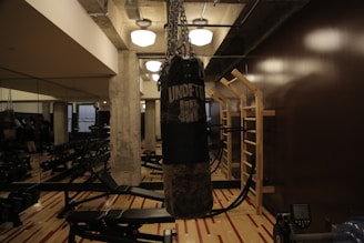 A dimly lit gym features a heavy punching bag hanging in the center, surrounded by workout benches and mirrored walls. The space has concrete pillars, wall bars, and exercise equipment lined up neatly. Overhead lights provide a warm, ambient glow.