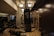 A dimly lit gym features a heavy punching bag hanging in the center, surrounded by workout benches and mirrored walls. The space has concrete pillars, wall bars, and exercise equipment lined up neatly. Overhead lights provide a warm, ambient glow.