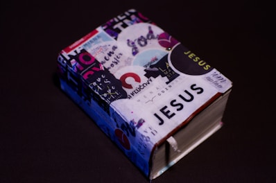 A small, thick book covered with various colorful stickers and texts, prominently featuring the word 'JESUS'. It is set against a plain dark background, enhancing the contrast of the vibrant collage on the book cover.