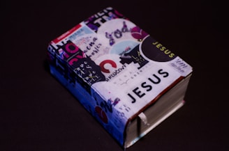 A small, thick book covered with various colorful stickers and texts, prominently featuring the word 'JESUS'. It is set against a plain dark background, enhancing the contrast of the vibrant collage on the book cover.