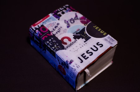 A small, thick book covered with various colorful stickers and texts, prominently featuring the word 'JESUS'. It is set against a plain dark background, enhancing the contrast of the vibrant collage on the book cover.