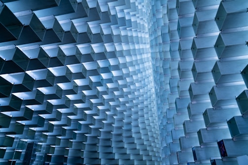 An array of geometric, modular blocks creates a visually intricate and repetitive pattern. The structure consists of metallic, rectangular shapes intersecting and forming a wave-like motion. These blocks are arranged meticulously to form a uniquely architectural and artistic space, with light reflecting off their surfaces to enhance the dimensional effect.