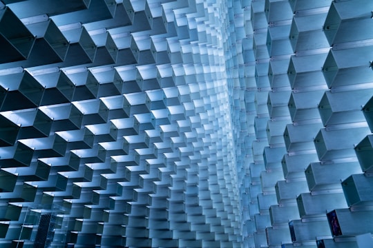 An array of geometric, modular blocks creates a visually intricate and repetitive pattern. The structure consists of metallic, rectangular shapes intersecting and forming a wave-like motion. These blocks are arranged meticulously to form a uniquely architectural and artistic space, with light reflecting off their surfaces to enhance the dimensional effect.