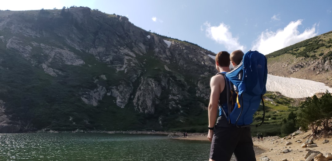 man carrying baby on blue backpack carrier facing mountain near body of water