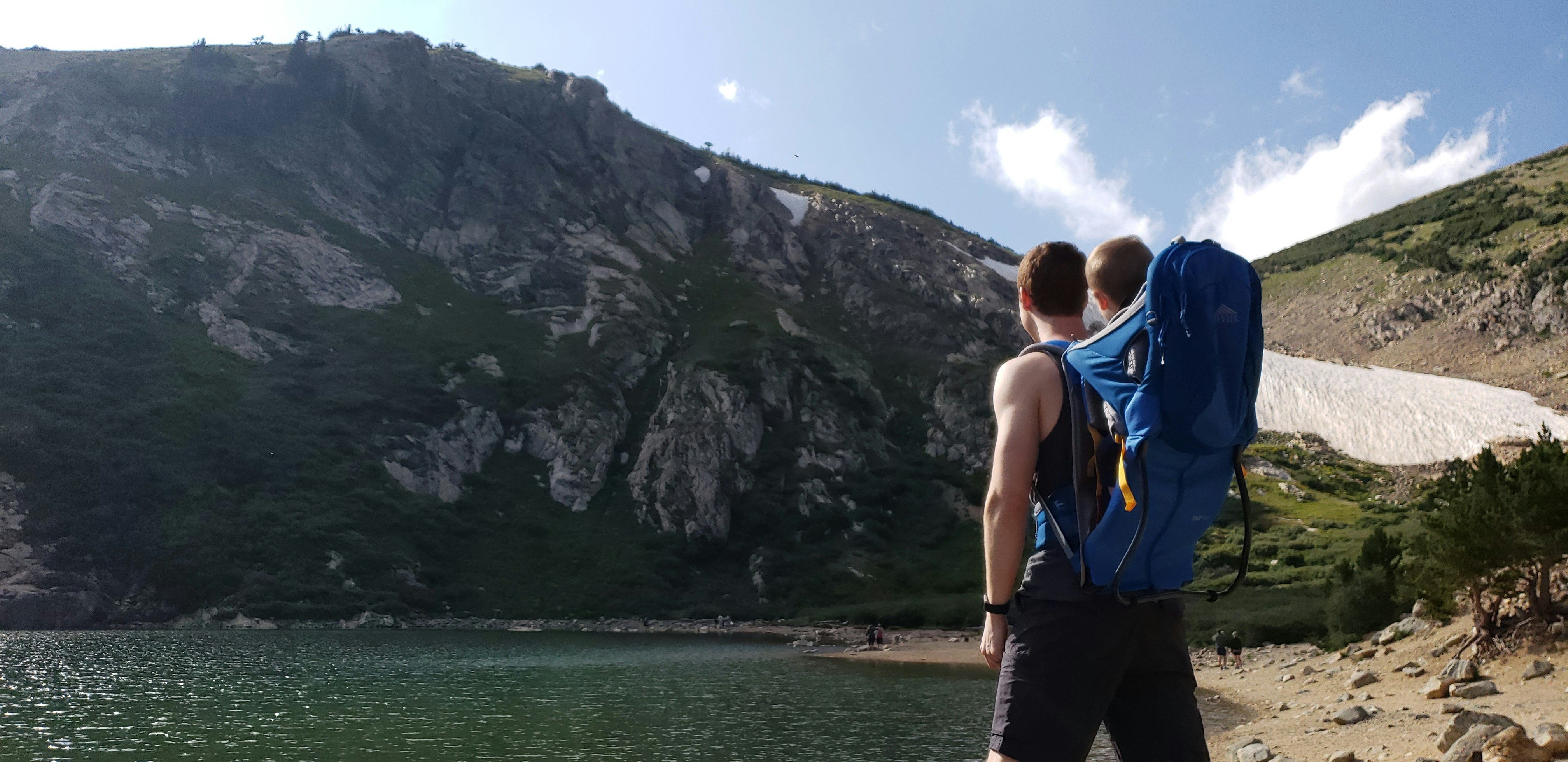 man carrying baby on blue backpack carrier facing mountain near body of water