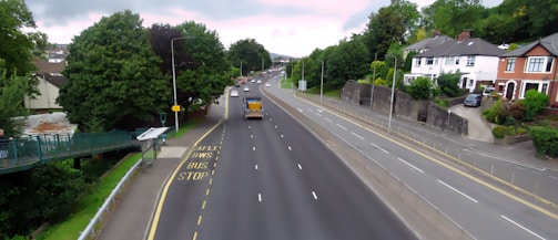 A well-maintained road with multiple lanes is lined with trees and residential houses. Traffic is moderate, with a few vehicles visible, including a truck. A pedestrian bridge is present on the left side, leading to a bus stop marked with yellow text on the road. The sky is partly cloudy, and the area appears suburban.