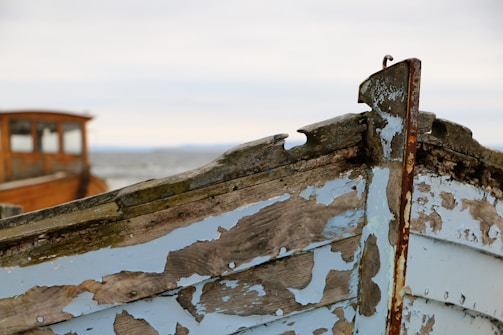 An old, weathered wooden boat with peeling blue and brown paint is shown. The wood appears rustic and aged, indicating it has been exposed to the elements for a significant time. In the blurred background, another wooden structure is visible, possibly part of another boat.