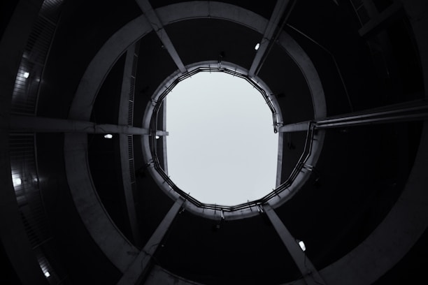 A circular structure with multiple levels is viewed from the bottom, creating a tunnel-like effect towards the sky. The architectural design features concrete elements with visible lighting fixtures around the edges.