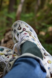 A pair of white canvas shoes with colorful character graphics, worn with blue jeans and green socks. The background consists of blurred greenery and brown tree trunks, suggesting a natural, outdoor setting.