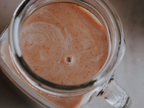 A top-down view of a glass jar filled with a creamy, light brown liquid that features subtle swirls and specks of darker brown. The jar's handle and edges are partially visible, creating a rustic appearance.