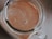 A top-down view of a glass jar filled with a creamy, light brown liquid that features subtle swirls and specks of darker brown. The jar's handle and edges are partially visible, creating a rustic appearance.