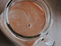A top-down view of a glass jar filled with a creamy, light brown liquid that features subtle swirls and specks of darker brown. The jar's handle and edges are partially visible, creating a rustic appearance.
