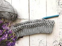 A crochet project in progress is displayed on a textured white wooden surface. There is a ball of gray yarn with a metallic sheen on the left side, accompanied by a teal-handled crochet hook inserted into the piece. Purple flowers are partially visible on the left.