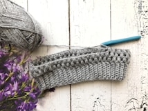 A crochet project in progress is displayed on a textured white wooden surface. There is a ball of gray yarn with a metallic sheen on the left side, accompanied by a teal-handled crochet hook inserted into the piece. Purple flowers are partially visible on the left.