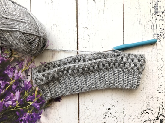 A crochet project in progress is displayed on a textured white wooden surface. There is a ball of gray yarn with a metallic sheen on the left side, accompanied by a teal-handled crochet hook inserted into the piece. Purple flowers are partially visible on the left.