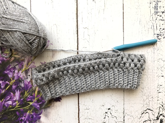 A crochet project in progress is displayed on a textured white wooden surface. There is a ball of gray yarn with a metallic sheen on the left side, accompanied by a teal-handled crochet hook inserted into the piece. Purple flowers are partially visible on the left.