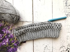 A crochet project in progress is displayed on a textured white wooden surface. There is a ball of gray yarn with a metallic sheen on the left side, accompanied by a teal-handled crochet hook inserted into the piece. Purple flowers are partially visible on the left.