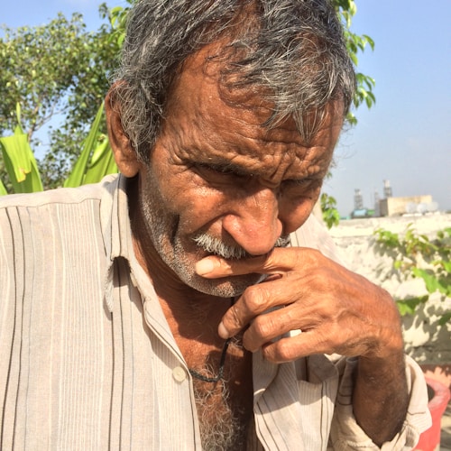 An elderly man with a thoughtful expression is outdoors in a bright setting. He is resting his hand on his chin and is wearing a striped shirt. The background includes green foliage and a clear blue sky.