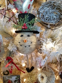A festive Christmas decoration featuring a handcrafted snowman ornament with a textured surface, adorned with a top hat, red berries, and green foliage. The snowman is nestled among white branches and surrounded by other decorative elements, including a woven ball resembling a bird's nest and red ribbons. Warm lighting enhances the holiday ambiance.