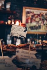 Several slices of bread, possibly toast, are displayed in a basket with a small sign in front. The sign contains text suggesting the food is a gluten-free option, describing it as 'Deluxe Choco-Amande.' The background includes blurred images, likely part of a cafe or coffee shop setting, with warm lighting adding to the cozy atmosphere.