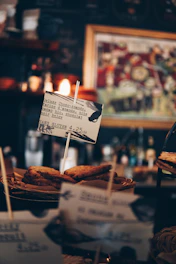 Several slices of bread, possibly toast, are displayed in a basket with a small sign in front. The sign contains text suggesting the food is a gluten-free option, describing it as 'Deluxe Choco-Amande.' The background includes blurred images, likely part of a cafe or coffee shop setting, with warm lighting adding to the cozy atmosphere.