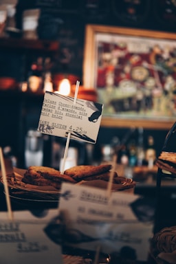 Several slices of bread, possibly toast, are displayed in a basket with a small sign in front. The sign contains text suggesting the food is a gluten-free option, describing it as 'Deluxe Choco-Amande.' The background includes blurred images, likely part of a cafe or coffee shop setting, with warm lighting adding to the cozy atmosphere.
