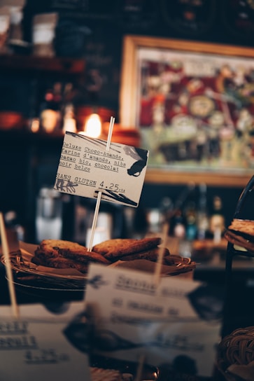 Several slices of bread, possibly toast, are displayed in a basket with a small sign in front. The sign contains text suggesting the food is a gluten-free option, describing it as 'Deluxe Choco-Amande.' The background includes blurred images, likely part of a cafe or coffee shop setting, with warm lighting adding to the cozy atmosphere.