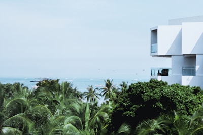 Lush green palm trees in the foreground frame a modern, minimalist building with large glass balconies on the right. In the background, a bright blue ocean extends to the horizon with several small boats dotting the water under a clear sky.