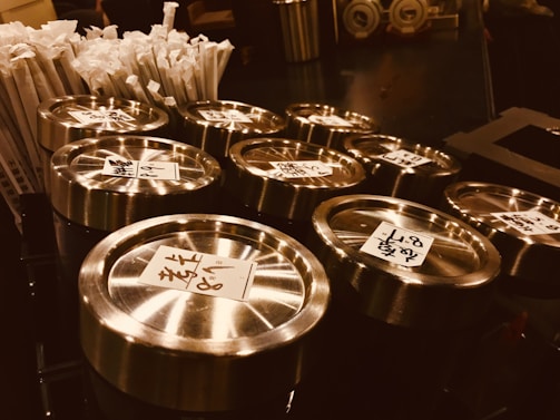 Several metal tins with glass lids and labels in a foreign script are placed closely on a countertop. To the left, there is a bundle of wrapped straws. The setting appears to be a counter in a café or restaurant.