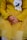 A child wearing a mustard yellow ribbed sweater and skirt sits with their hands gently clasped. The outfit features an embroidered patch showing a sleeping face with closed eyes and a bow detail, alongside a lace accent.