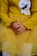 A child wearing a mustard yellow ribbed sweater and skirt sits with their hands gently clasped. The outfit features an embroidered patch showing a sleeping face with closed eyes and a bow detail, alongside a lace accent.