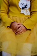 A child wearing a mustard yellow ribbed sweater and skirt sits with their hands gently clasped. The outfit features an embroidered patch showing a sleeping face with closed eyes and a bow detail, alongside a lace accent.