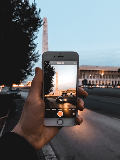 A hand holds a smartphone capturing an image of a tall obelisk-like structure against a cityscape backdrop during twilight. The smartphone screen displays a more vibrant, warmer-toned version of the scene with a road and building also visible.