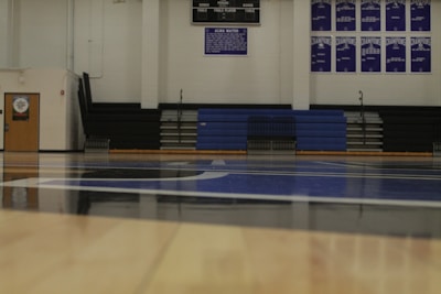 An indoor gymnasium with wooden flooring, blue and white court markings, and empty bleachers in the background. There are banners and a scoreboard on the wall, and a door to the left side.
