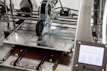 A 3D printer in operation, with a close-up view of the printing head moving over the print bed. The printer's mechanical components and electronic controls are visible, emphasizing precision and technology.