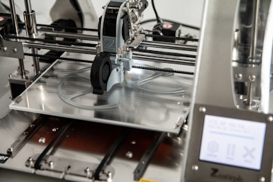 A 3D printer in operation, with a close-up view of the printing head moving over the print bed. The printer's mechanical components and electronic controls are visible, emphasizing precision and technology.