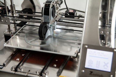 A 3D printer in operation, with a close-up view of the printing head moving over the print bed. The printer's mechanical components and electronic controls are visible, emphasizing precision and technology.