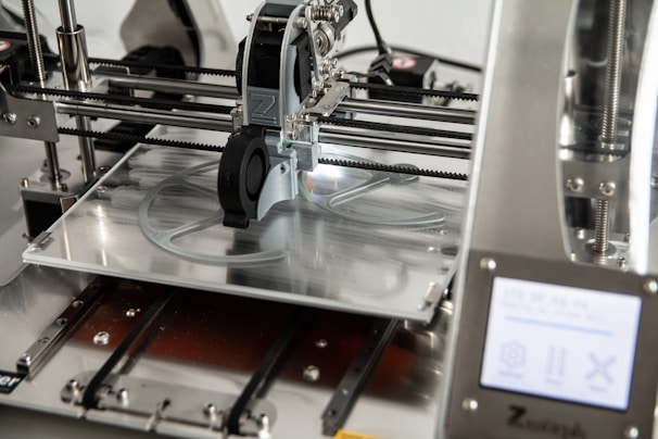 A 3D printer in operation, with a close-up view of the printing head moving over the print bed. The printer's mechanical components and electronic controls are visible, emphasizing precision and technology.