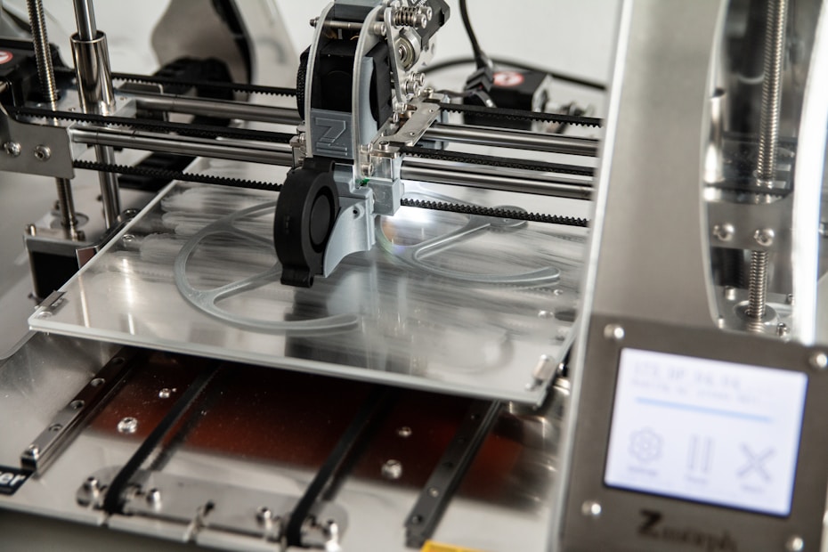 A 3D printer in operation, with a close-up view of the printing head moving over the print bed. The printer's mechanical components and electronic controls are visible, emphasizing precision and technology.