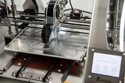A 3D printer in operation, with a close-up view of the printing head moving over the print bed. The printer's mechanical components and electronic controls are visible, emphasizing precision and technology.
