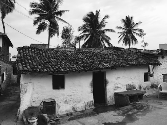 A rustic house with a clay-tiled roof situated in a rural setting, surrounded by towering palm trees. The structure appears weathered, with a sturdy white exterior and a few small windows. A large water barrel and some household items are placed outside, alongside a plastic chair, suggesting daily life activities. The dirt path leading to the house is visible, emphasizing its simplicity and charm.