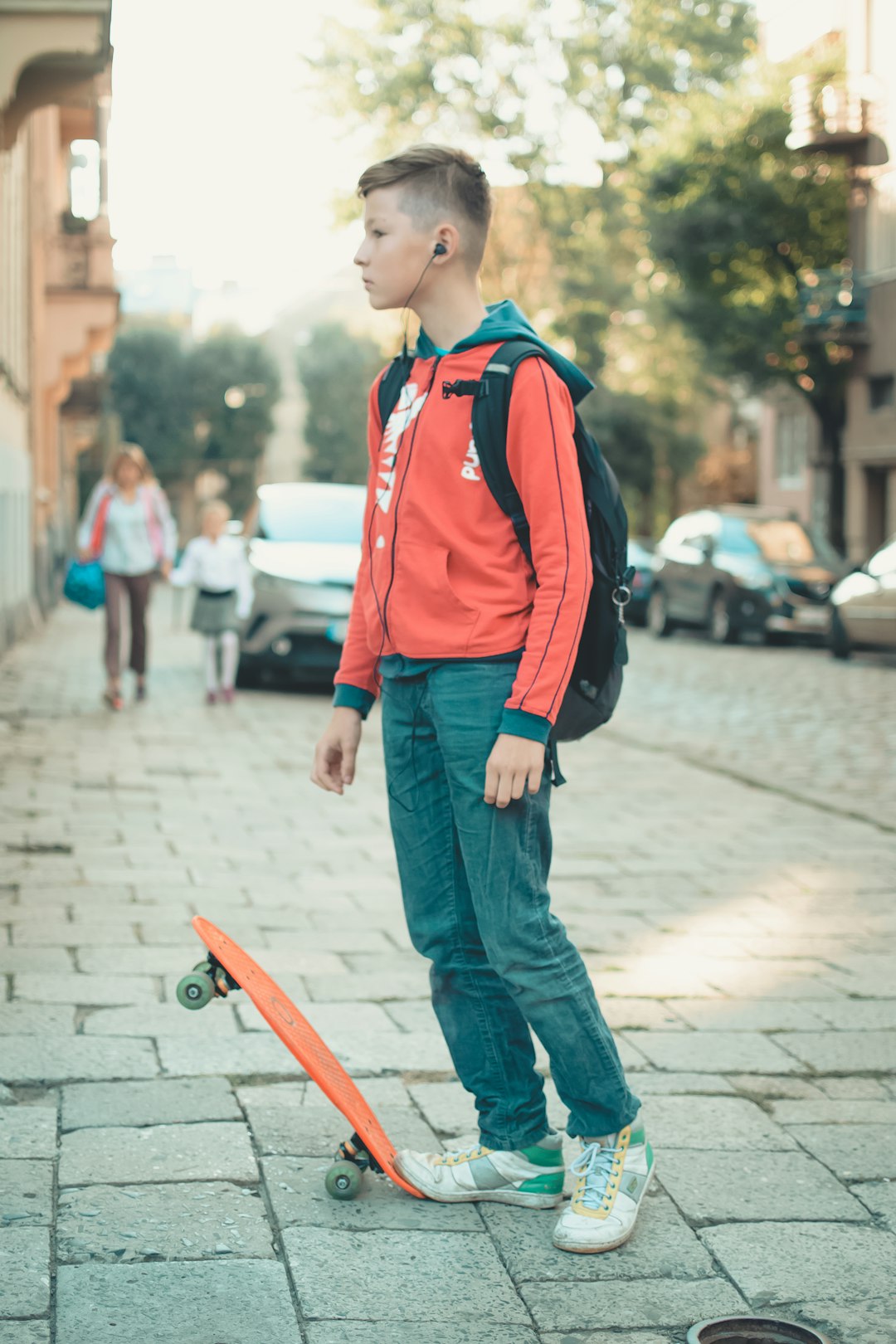 boy with backpack and earbuds standing near orange skateboard