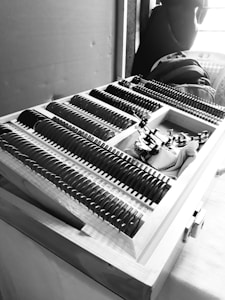 A black and white image featuring a wooden box filled with a neatly organized assortment of circular lenses or glass pieces. Each piece has markings or measurements beside it, possibly representing different lens strengths or sizes. The box appears to be part of a larger setup, possibly indicating an optical or measurement-related task. In the background, several items like a backpack and a jacket are partially visible.