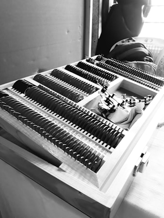 A black and white image featuring a wooden box filled with a neatly organized assortment of circular lenses or glass pieces. Each piece has markings or measurements beside it, possibly representing different lens strengths or sizes. The box appears to be part of a larger setup, possibly indicating an optical or measurement-related task. In the background, several items like a backpack and a jacket are partially visible.