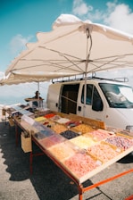 A roadside market stall features a variety of colorful dried fruits displayed in trays. The stall is set up next to a white van and shaded by a large umbrella. The background showcases a clear blue sky with a few clouds, suggesting a sunny day.